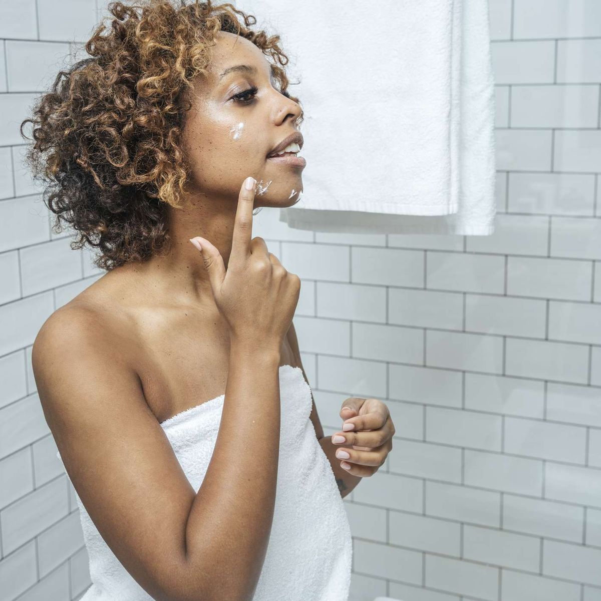 אישה מורחת קרם פנים בחדר אמבטיה לבן עם אריחים
