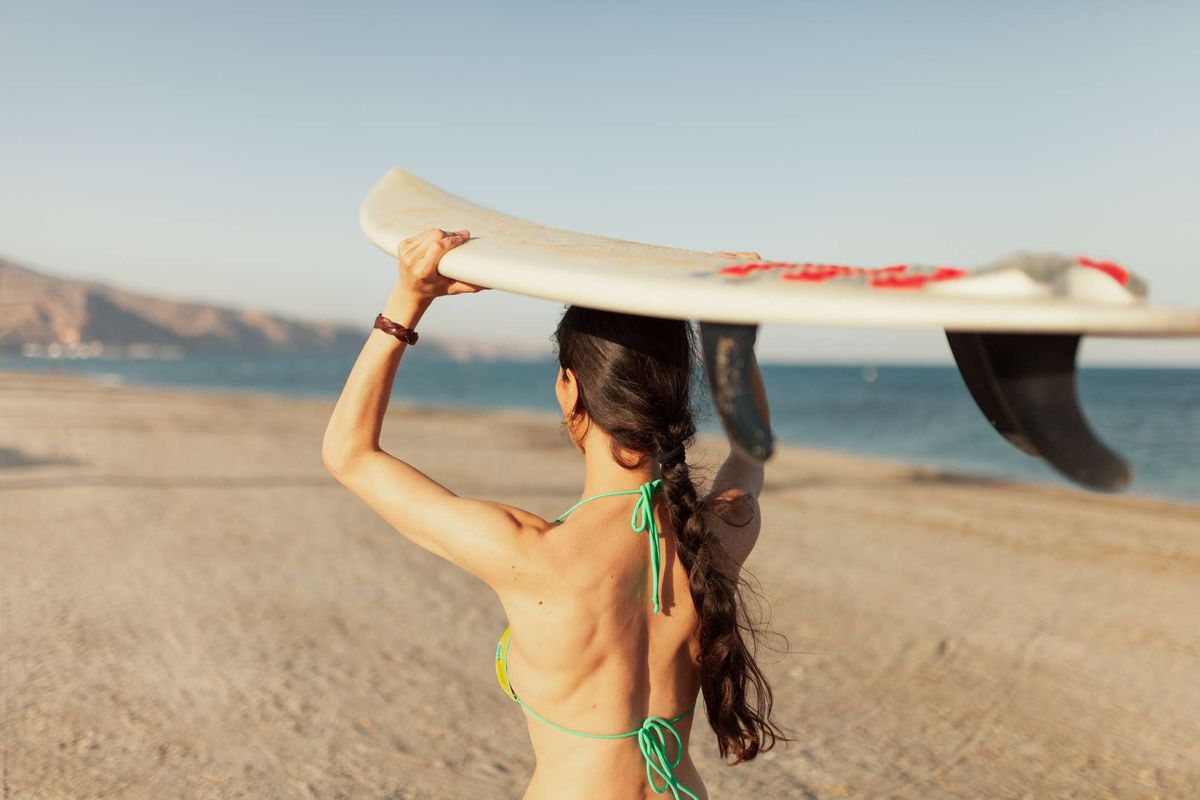 Žena koja hoda plažom noseći dasku za surfanje naslonjenu na glavu