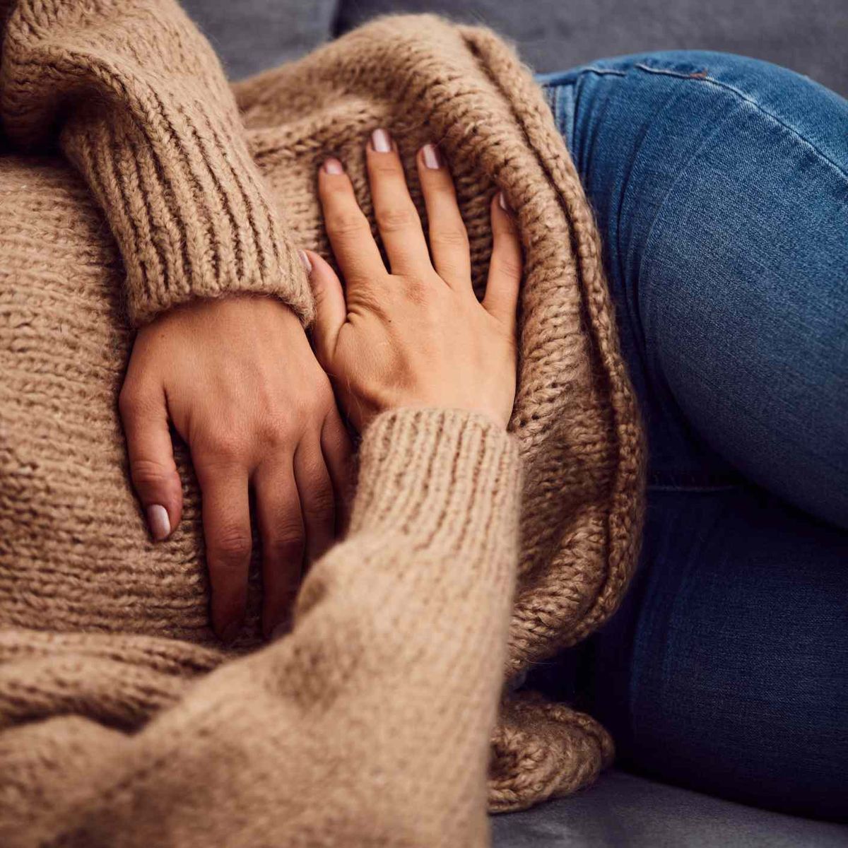 Frau in einem braunen Pullover umklammert ihren Bauch und liegt auf einer Couch