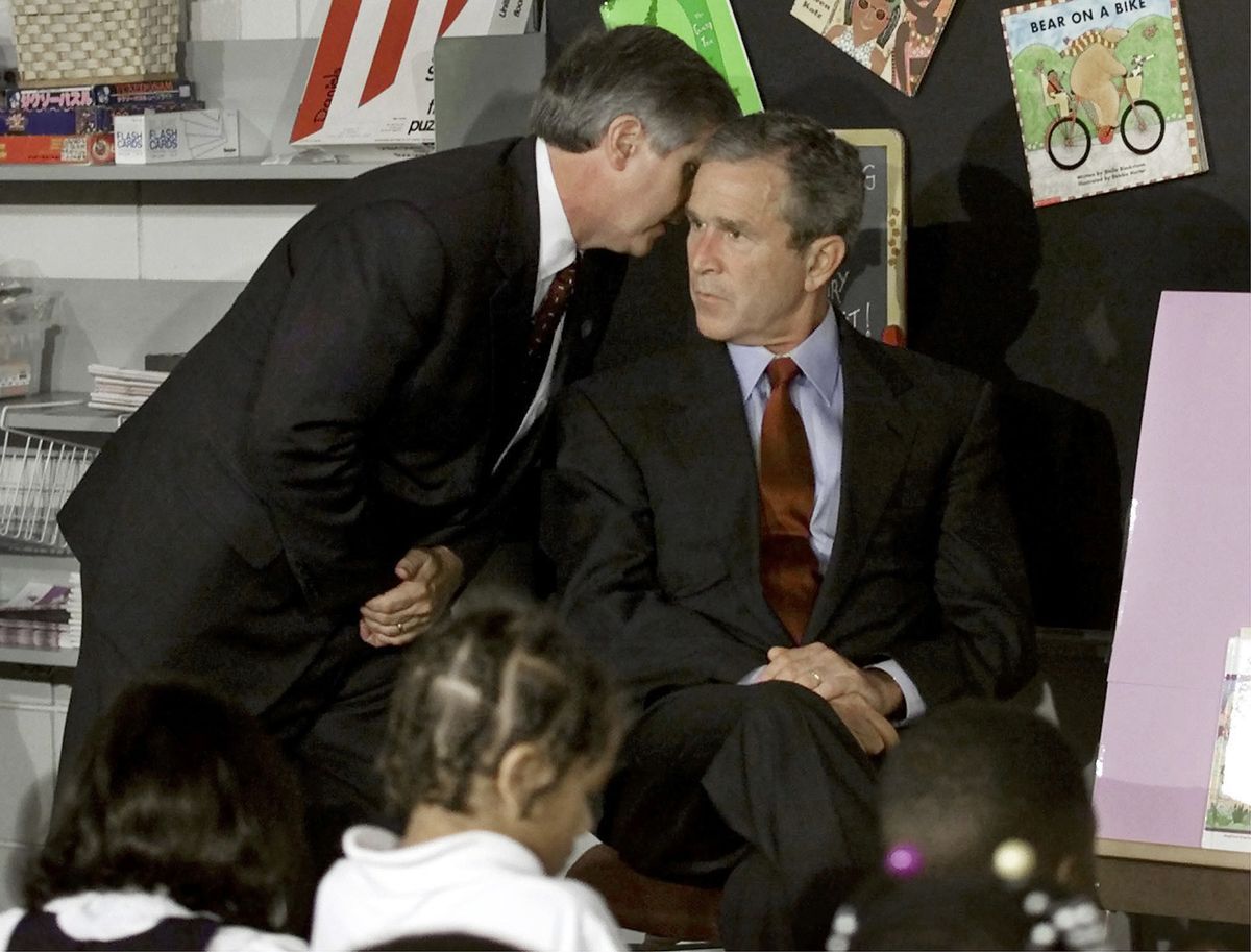 Президент Джордж Буш өте байсалды көрінеді, ал костюм киген басқа адам оның құлағына сыбырлайды