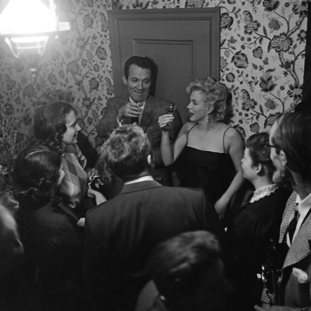 Marilyn Monroe hält ein kleines Glas Alkohol hoch, umgeben von Leuten auf einer Party