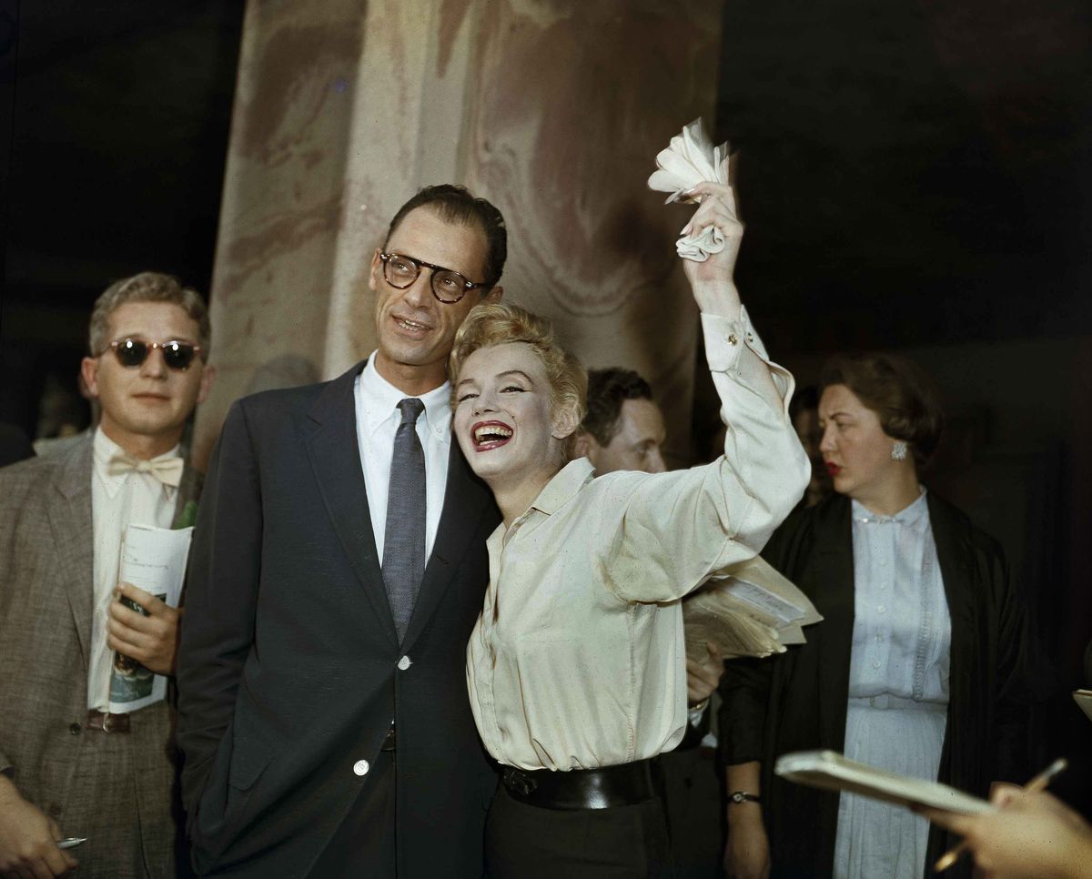 Marilyn Monroe lächelt breit und schwenkt ihre Handschuhe in einer Hand, als sie neben ihrem Ehemann Arthur Miller steht
