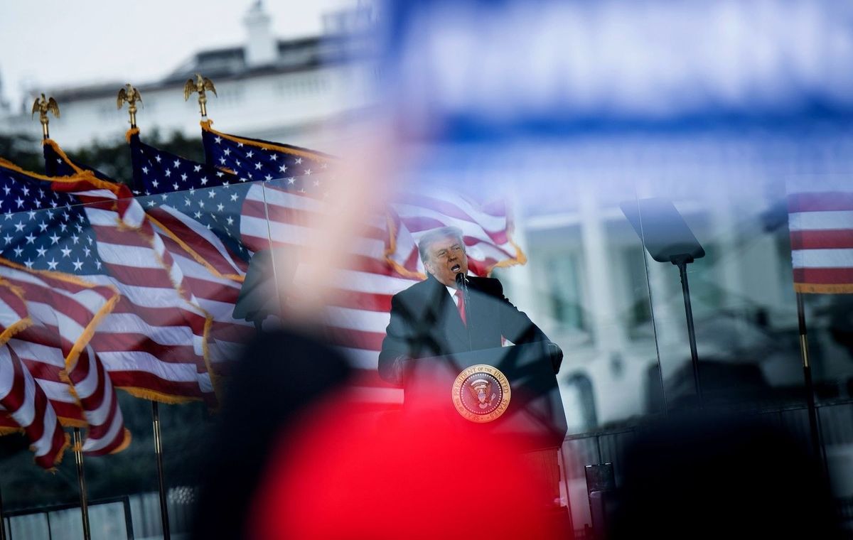 Trump se para detrás de un atril con un sello presidencial, frente a banderas estadounidenses ondeando al viento; En primer plano, los seguidores desenfocados ondean carteles y banderas.